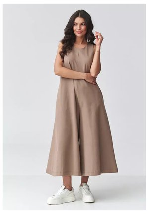 Kobieta z długimi ciemnymi włosami, ubrana w bezrękawnikowy, beżowy kombinezon z szerokimi nogawkami oraz białe sneakersy na platformie, stojąca na tle jednolitego tła.