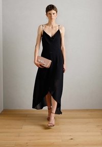 Robe noire avec un décolleté drapé, un ourlet asymétrique et des fines bretelles avec des accents en métal. Associée à une pochette rose et des talons strappés.