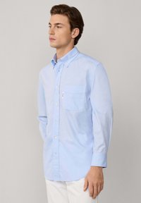 Camisa abotonada de color azul claro, hecha de algodón, con cuello, un bolsillo en el pecho y mangas largas. Textura suave con sutiles detalles de costura.