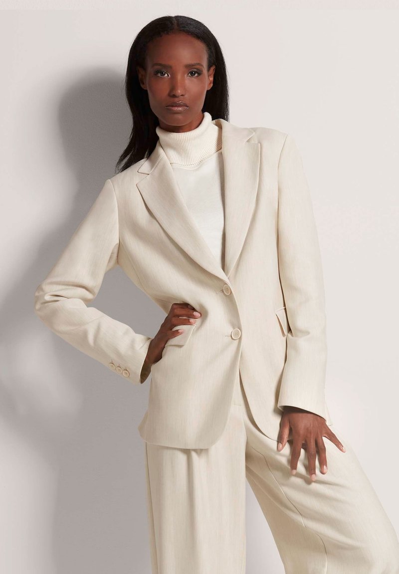 Crème-kleurige op maat gemaakte blazer met een reverskraag, twee knopen en subtiele verticale strepen, gecombineerd met wijde broekspijpen.