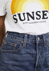 T-shirt blanc avec un graphique de coucher de soleil jaune et orange ; jeans en denim bleu taille haute avec couture visible et fermeture à bouton en métal.