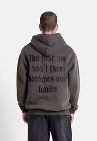 Man i mörk hoodie med texten "Rädslan vi inte möter blir våra begränsningar" på baksidan, står mot en enkel ljus bakgrund.