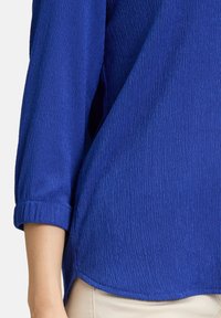 Haut texturé bleu avec des manches trois-quarts, présentant un motif légèrement irrégulier et un ourlet décontracté. Le tissu semble léger et respirant.