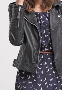 Musta nahkamoottoripyörätakki, jossa on tikatut olkaosuudet, hopeiset vetoketjut ja vyölliset yksityiskohdat, puettuna kuviolaukkujen päälle.