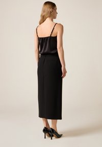 Camisola de satén negra con tirantes finos, combinada con una falda lápiz negra de cintura alta. La modelo lleva tacones de charol y pendientes llamativos.