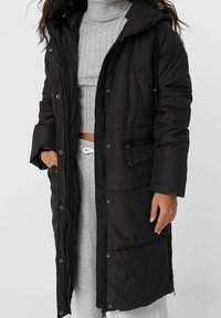 Sort puffermantel med lang design, der har en lynlåslukning, trykknapper, sidelommer og en hætte. Materialet ser tekstureret og varmt ud.