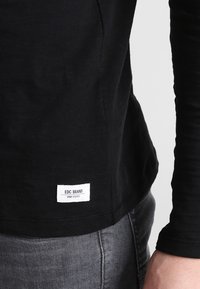 Czarna koszula z długim rękawem, uszyta z miękkiego materiału, z okrągłym dekoltem i metką na dolnym szwie z napisem "EDC BRAND DNM GOODS" na białym tle.