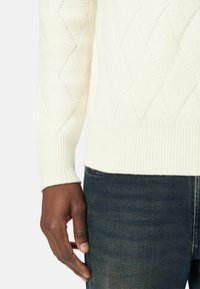 Crème-kleurige kabelgebreide trui met een gestructureerd ruitpatroon, geribbelde manchetten en zoom. Gecombineerd met donkerdenim jeans.