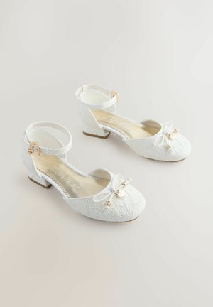 Hvide balletsko med tekstureret stof, ankelrem, sløjfedetalje og lille, solid hæl. Gyldent hardware-detalje nær sløjfen.