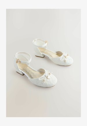 Witte ballet schoentjes met een gestructureerde stof, enkelbandje, strikaccent en kleine, stevige hak. Gouden hardware detail nabij de strik.