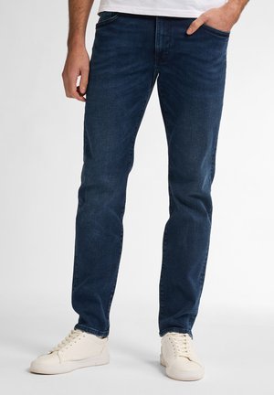 Dark blue denim jeans with a slim fit, featuring front pockets, a zipped fly, and a slight fade in the fabric texture. Pair of white sneakers.