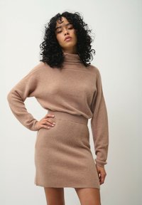 Beżowy sweter robiony na drutach z wysokim kołnierzem i długimi rękawami, w połączeniu z dopasowaną, prążkowaną spódnicą, tworzącą spójną, teksturowaną stylizację.