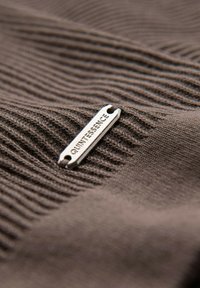 Tessuto marrone con texture e righe verticali, con un'etichetta in metallo silver con testo in rilievo "QUINTESSENCE" attaccata.