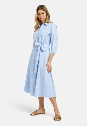 Woman standing wearing a blue and white vertical striped midi shirt dress with a tied waist belt and tan flat sandals.