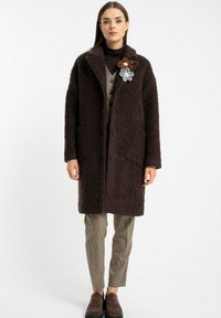 Brązowy futrzany płaszcz z dużymi klapami i guzikami, w połączeniu z w kratkę kamizelką, ciasno dopasowanymi spodniami w kratkę i ciemnymi butami. Akcent w postaci broszki w kształcie kwiatu.