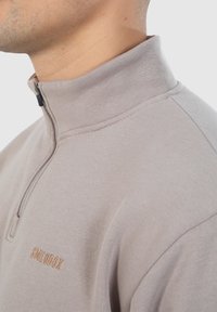 Lichtbeige half-zip pullover gemaakt van een gladde stof, met een subtiele textuur en een geborduurd logo in een donkerdere tint.