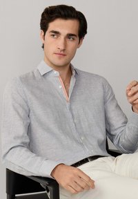 Mladić sjedi i nosi tanku košulju s dugim rukavima i prugama, svijetlih hlača, gledajući blago u stranu.