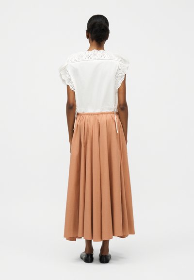 Γυναίκα στέκεται με την πλάτη γυρισμένη, φορώντας λευκή μπλούζα χωρίς μανίκια με σχέδιο eyelet και μακριά πλισέ καφέ φούστα με μαύρα φλατ παπούτσια σε λευκό φόντο.