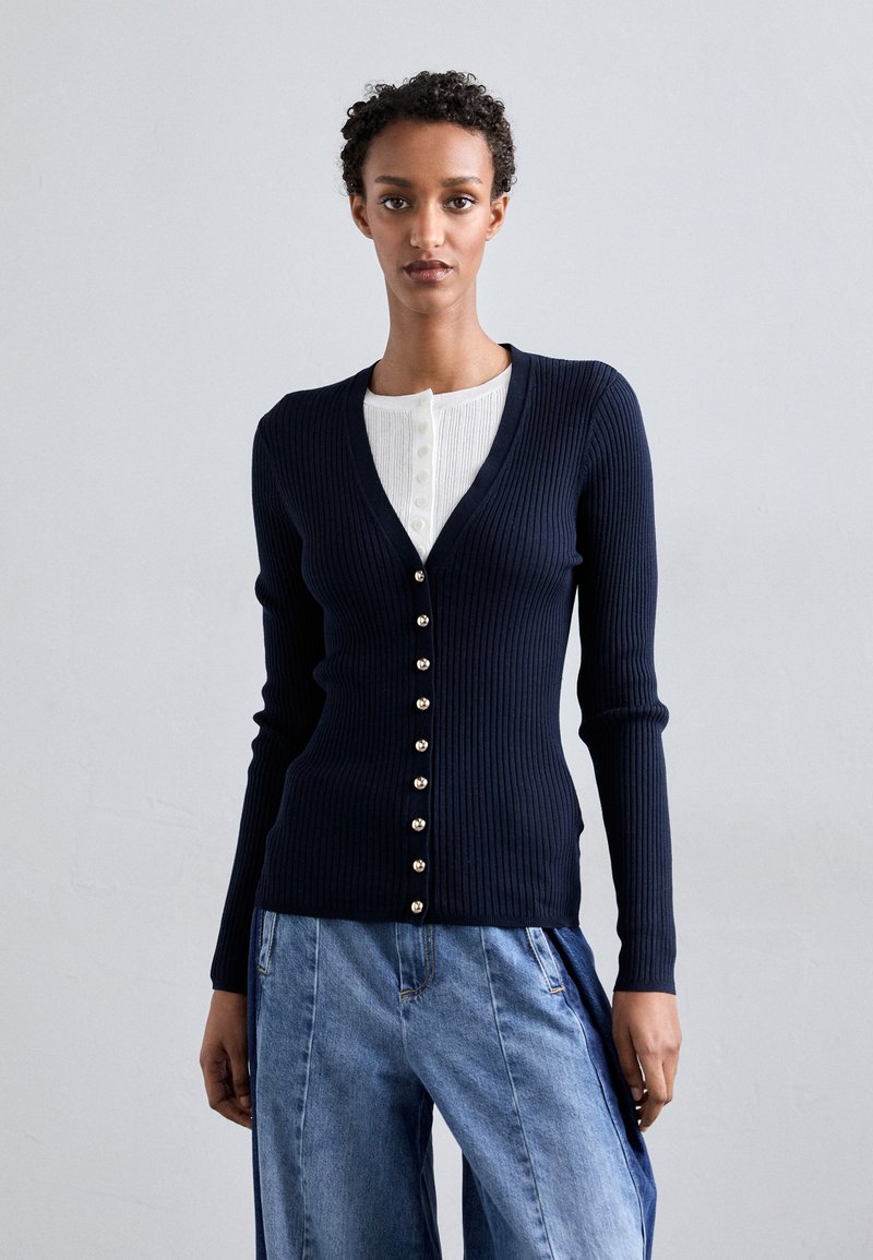 Marineblaue gerippte Strickjacke mit tiefem V-Ausschnitt, versehen mit goldfarbenen Knöpfen auf der Vorderseite. Kombiniert mit hellblauen Jeans.