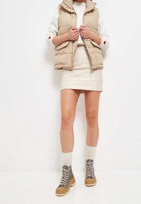 Beżowa pikowana kamizelka z zamkiem błyskawicznym, zestawiona z kremową pikowaną mini spódnicą. Szare botki sznurowane na grubej beżowej podeszwie. Widoczne kremowe skarpetki.