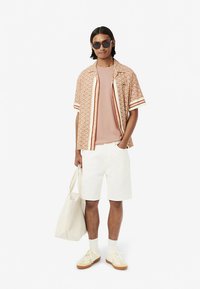 Camisa de manga curta em bege com padrão geométrico, t-shirt rosa claro, calções brancos, sapatilhas bege e uma bolsa ligeira.