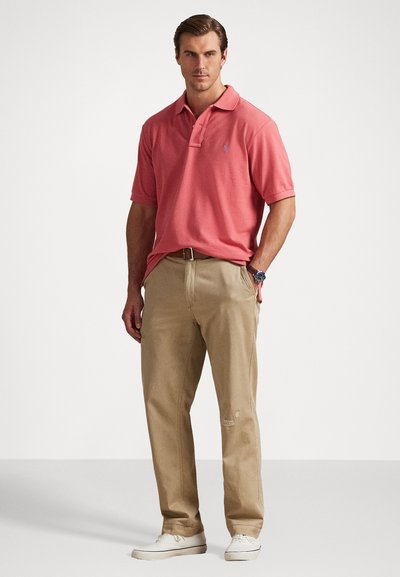 Κόκκινο πόλο μπλουζάκι με κλασικό γιακά, φορεμένο με μπεζ παντελόνια. Ρολόι στον καρπό και λευκά sneakers. Απλός, χαλαρός σχεδιασμός.