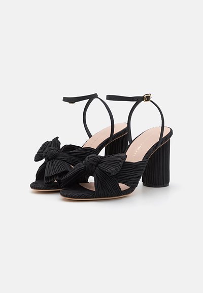 Sandalias de tacón alto negras con parte superior de tela plisada, detalle de lazo, puntera descubierta y correa de tobillo ajustable sobre un tacón grueso.