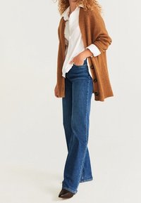 Brun strikket cardigan med store knapper, båret over en hvid skjorte og blå flared jeans. Outfit'et har en afslappet pasform og struktureret stof.