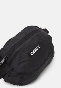Black nylon bag with a textured surface, featuring a top zipper and a rectangular "OBEY" logo patch on the front. Compact design.