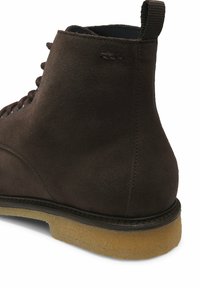 Brązowy zamszowy botek za kostkę z okrągłym noskiem, sznurowanym zapięciem i teksturowaną gumową podeszwą. Posiada pętelkę z tyłu oraz dekoracyjne przeszycia.