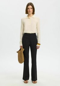 Femme portant un polo crème à manches longues, un pantalon noir taille haute, des tongs, tenant un sac marron texturé et portant des bracelets colorés au poignet.