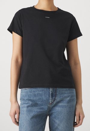 Vrouw draagt een effen zwart T-shirt met korte mouwen met klein wit "PINKO"-logo en lichtblauwe spijkerbroek tegen een witte achtergrond.