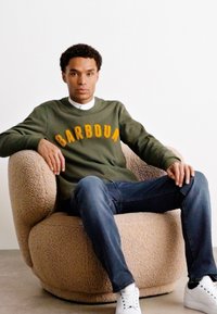 Olivově zelená mikina s nápisem "BARBOUR" žlutým písmem, spárovaná s modrými džíny a bílými teniskami, sedící na béžové texturované židli.