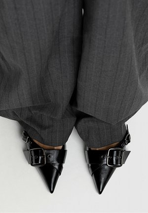 Czarne buty w szpic z szerokimi paskami zapinanymi na klamry, noszone z luźnymi, ciemnymi spodniami w drobną prążkę na jasnym tle.