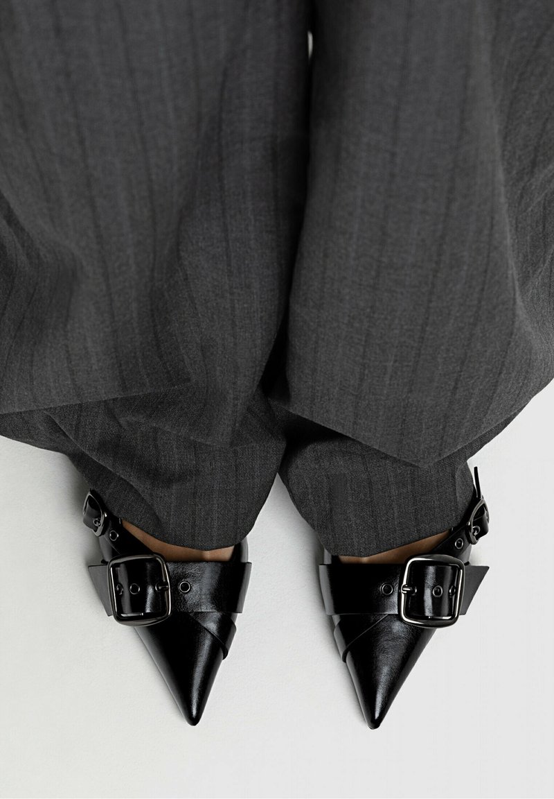 Chaussures noires à bout pointu avec larges sangles à boucle, portées avec un pantalon ample à fines rayures foncé sur un fond clair.