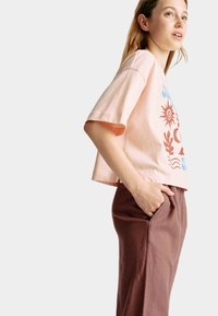 Korte roze katoenen t-shirt met een oceaan-thema graphic, gecombineerd met bruine comfortabele broek. Model staat met één hand in de zak.