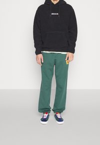 Felpa nera con cappuccio e tasca frontale con logo; pantaloni della tuta verdi con dettaglio di ricamo sottile; sneaker blu e bianche.