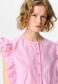 Blusa in cotone rosa con maniche arricciate e scollatura rotonda, con chiusura a bottoni sul davanti. Texture liscia e finitura morbida.