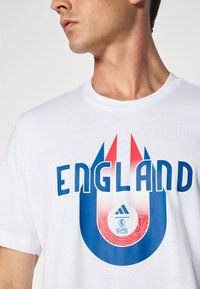 Hvid bomulds t-shirt med stor blå "ENG" tekst med røde og blå grafiske elementer samt Adidas-logoen med et UEFA Euro 2024 emblem.