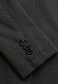 Manga de traje gris oscuro con tres botones negros etiquetados "OMBRE," mostrando una costura ordenada y textura de tela.