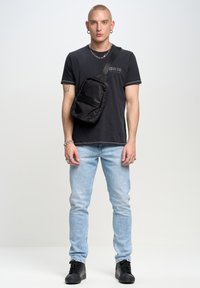 Bolso bandolera negro acolchado llevado en un hombro; cuenta con un bolsillo frontal con cremallera y una correa ajustable. El modelo lleva una camiseta negra y jeans claros.