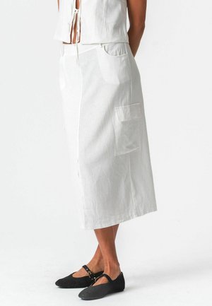 Vrouw draagt een witte midi-rok met zijzak en een wit mouwloos topje, gecombineerd met zwarte platte Mary Jane schoenen.