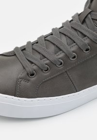 Zapatilla de cuero gris con cordones planos, ojetes de metal y una suela de goma blanca. Presenta costuras sutiles y una textura suave.