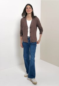 Gilet en tricot marron avec boutons, porté sur un haut blanc et un jean évasé bleu. Le modèle se tient devant un fond neutre avec des baskets claires.