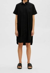 Svart skjortklänning med kort ärm, knäppning fram, avslappnad passform och sidofickor. Bärs med svarta halksandaler, platt sula.