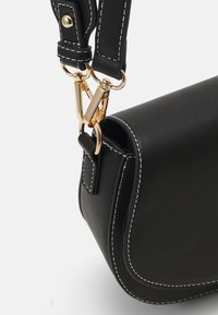 Musta nahkainen käsilaukku, jossa on valkoiset kontrastitikkaukset, kullanvärinen lukko ja sileä pinta sekä jäsennelty muoto.