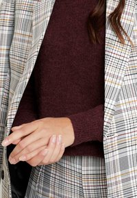 Suéter de punto color burdeos combinado con un blazer de cuadros que presenta colores blancos, negros y acentos otoñales. Primer plano de brazos cruzados.