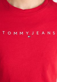Červené bavlněné tričko s nápisem "TOMMY JEANS" v bílé barvě nad malým modro-bílým logem, s žebrovaným kulatým výstřihem.