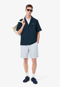 Ναυτικό κοντομάνικο πουκάμισο με ανάγλυφο σχέδιο, λευκό πόλο από κάτω, ριγέ ανοιχτό μπλέ σορτς και ναυτικά παπούτσια, κρατώντας μια μπεζ τσάντα.