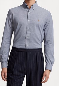 Vaaleanharmaasta kankaasta valmistettu, pitkähihainen kauluspaita, jossa pieni kirjailtu hevosen logo ja valkoiset napit. Yhdistetty tummiin raidallisiin housuihin.